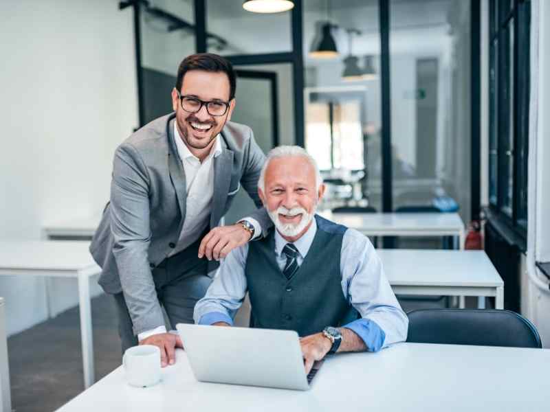 Empresario veterano y su sucesor joven sonriendo frente a una laptop en la oficina, representando la continuidad y liderazgo compartido en una empresa familiar.