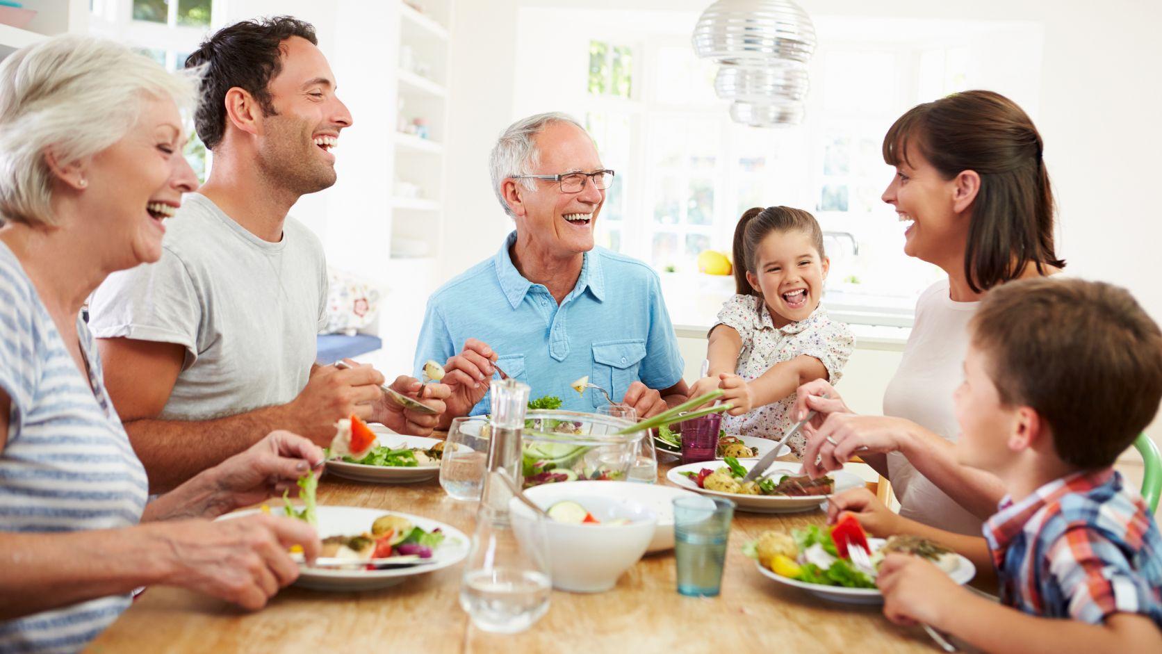 Familia de varias generaciones reunida alrededor de una mesa compartiendo alimentos durante una comida en casa.